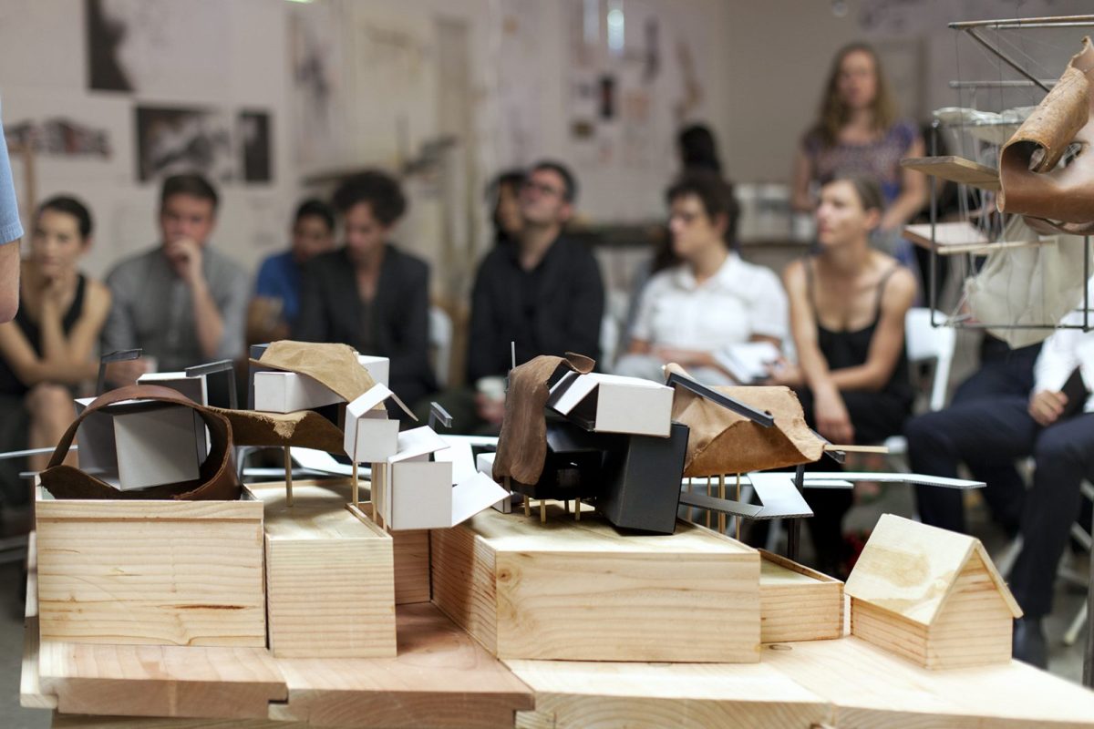 A Sculptural form of pinewood, white paper, and brown leather looking fabric is in the foreground with a group of intently watching students sitting in the background.