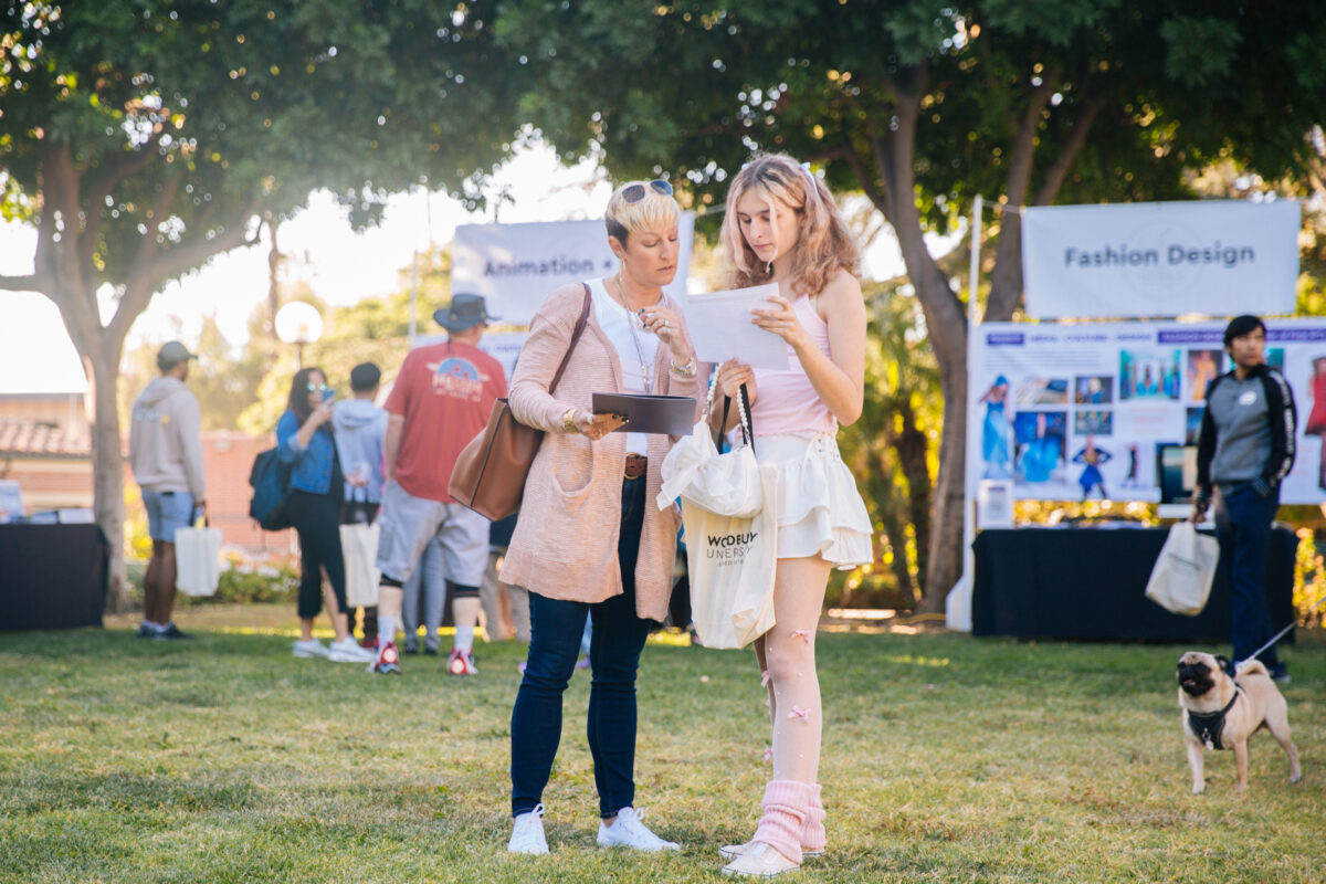 A student holding a piece of paper and their parent stand on the grass, both wear pink and have an intrigued look on their faces.