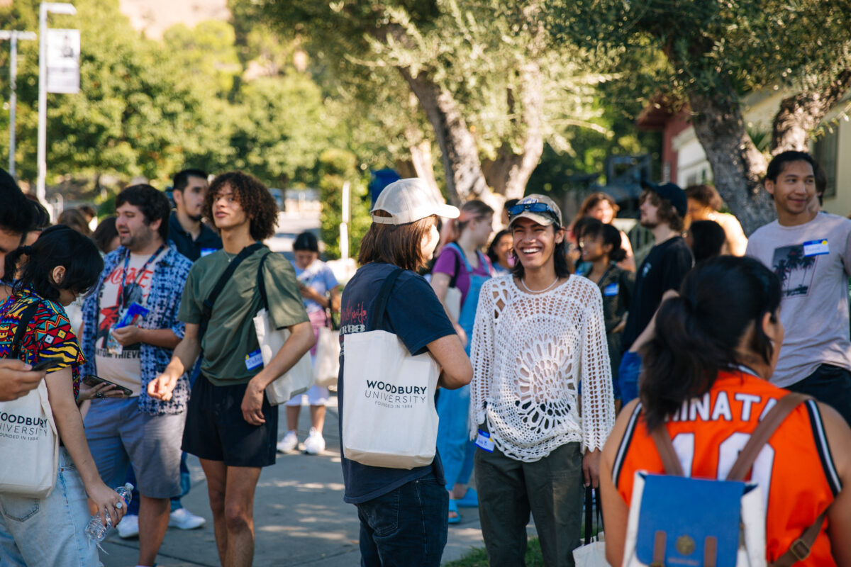 New students gathering in a crowd in the shade of oak trees. The central student has a wide smile and wears a white crochet shirt.